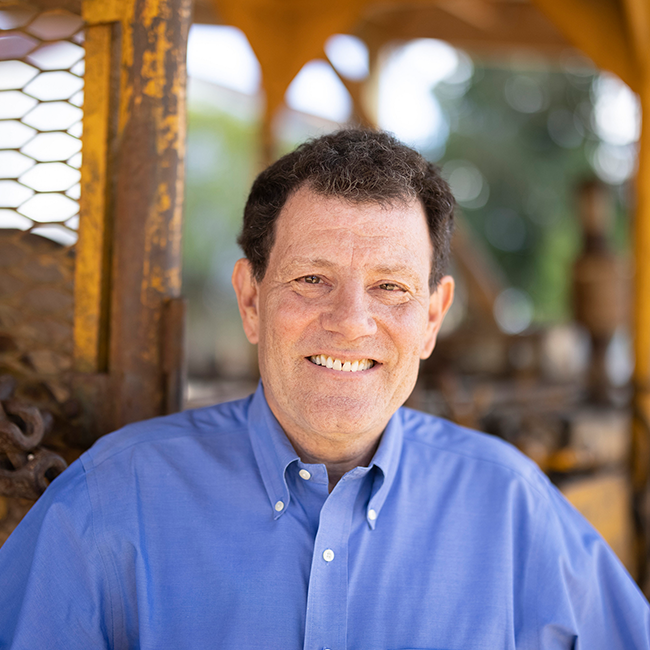 An image of a man named Nicholas Kristof. He has white skin, short brown hair, and is wearing a blue button-up shirt, smiling at the camera with an ambiguous outdoor background.