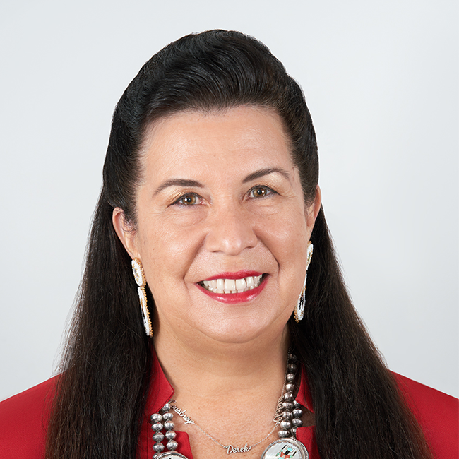 An image of a woman named Valerie Red-Horse Mohl. She has white skin, brown-ish eyes, and long dark brown hair. She is wearing long white earrings, a red shirt, and a decorative necklace and smiling at the camera against a white background.