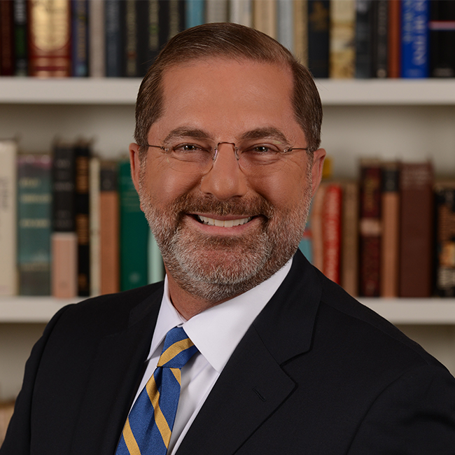 An image of a man named Alex Azar. He is standing in front of a bookshelf and smiling at the camera. He is middle-aged in appearance, has white skin, short brown hair, and a gray-ish brown stubbled beard. He is wearing a suit jacket over a white collared shirt with a blue and yellow striped tie, and round glasses.