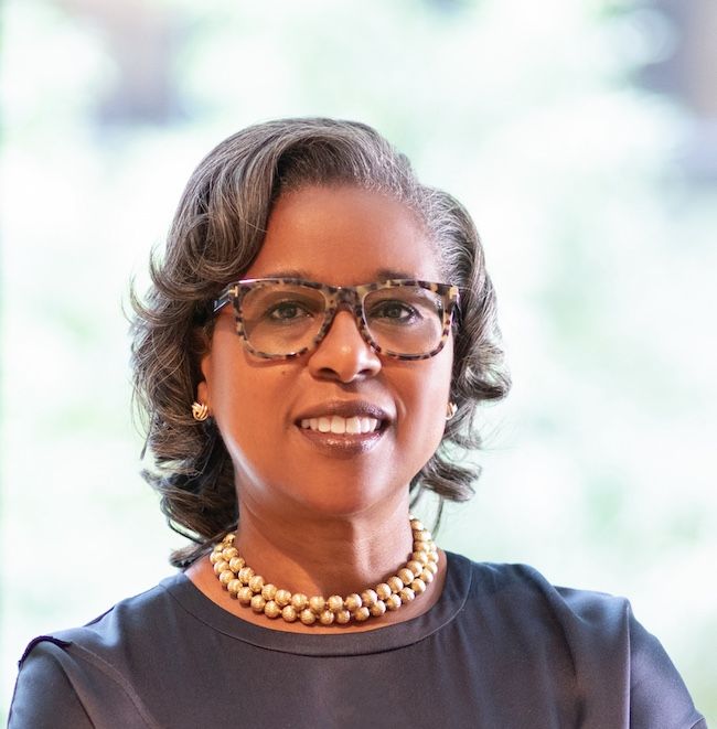 Professional headshot of a woman with short, styled gray hair wearing tortoiseshell glasses, a dark top, gold earrings, and a double-strand gold beaded necklace. She is smiling softly, with a blurred natural background behind her.