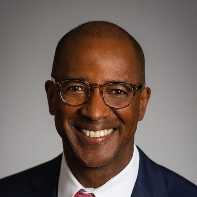 An image of a man named Eric Stevenson. He has brown skin, and shaved dark hair. He is wearing glasses and a suit and smiling at the camera against a gray background.