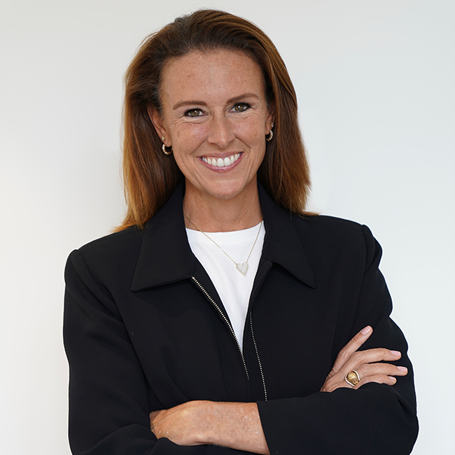 An image of a woman named Jill Popelka, smiling at the camera with her arms crossed against a white background. She has gold hoop earrings, a dark blue jacket over a white shirt, and has shoulder-length brunette hair and white skin.