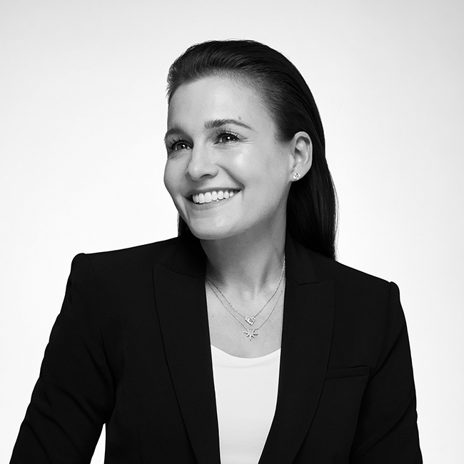 An black and white headshot of a woman named Jordana Upton. She appears to have white skin, long dark hair, and she is wearing a dark blazer over a white shirt while smiling at the camera against a white background.