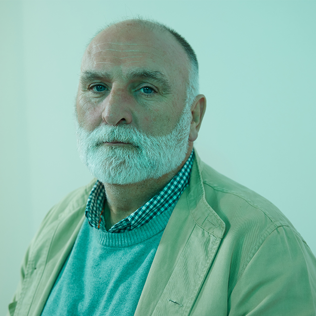 An image of a man named José Andrés. He is standing against a light green-blue background and staring seriously at the camera. He is older in appearance with a white beard, bald head, blue-ish eyes, and he is wearing a tan jacket over a sweater over a collared plaid shirt.