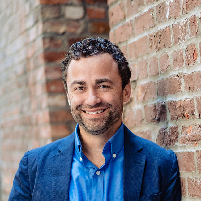 A headshot of a man named Matt Oppenheimer. He is standing against a brick wall and smiling at the camera. He has white skin, short brown hair, and a slight beard, and he is wearing a blue blazer with a blue collared shirt.