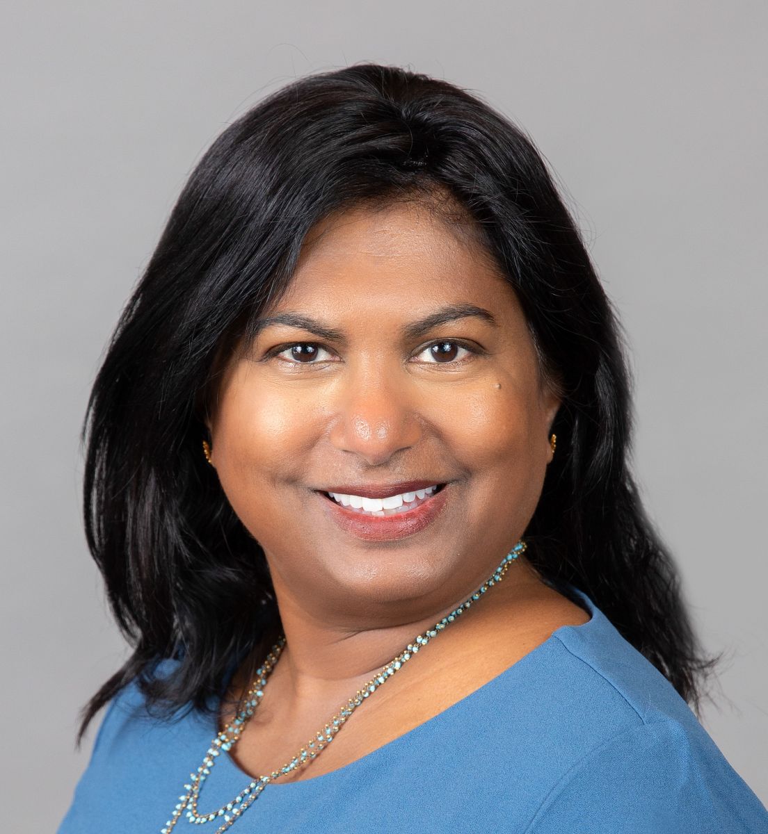 An image of a female-presenting person who is Indian in appearance. She is smiling at the camera and standing against a gray background. She has long black hair, tan skin, brown eyes, and is wearing a light blue shirt with a long necklace.