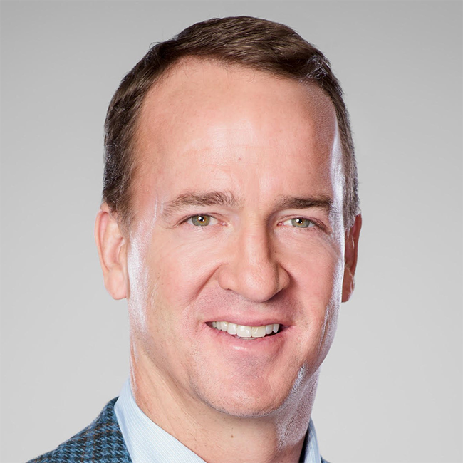 A headshot of Peyton Manning. He has white skin, short brown hair, and a blue suit and is smiling at the camera against a gray background.