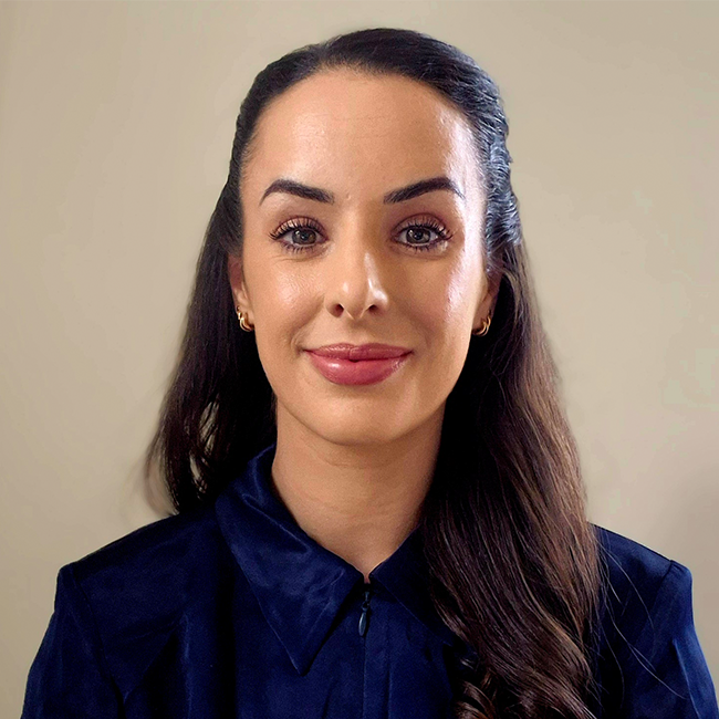A headshot of a woman named Rose Prinelle, who is standing against a beige background and slightly smiling at the camera. She has long dark brunette hair, white skin, and is wearing a dark blue shirt.