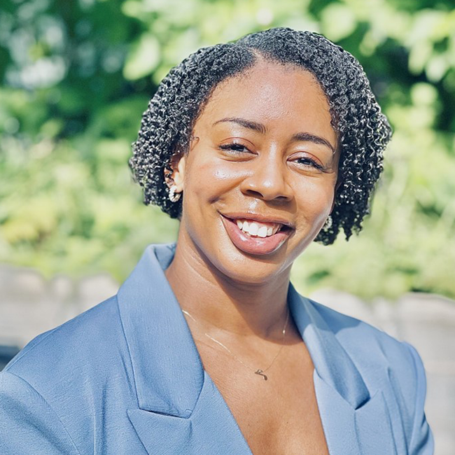 An image of a woman named Jasmine Jones. She has short black curly hair, brown skin, and brown eyes. She is smiling at the camera against an outdoor background and wearing a light blue blazer.
