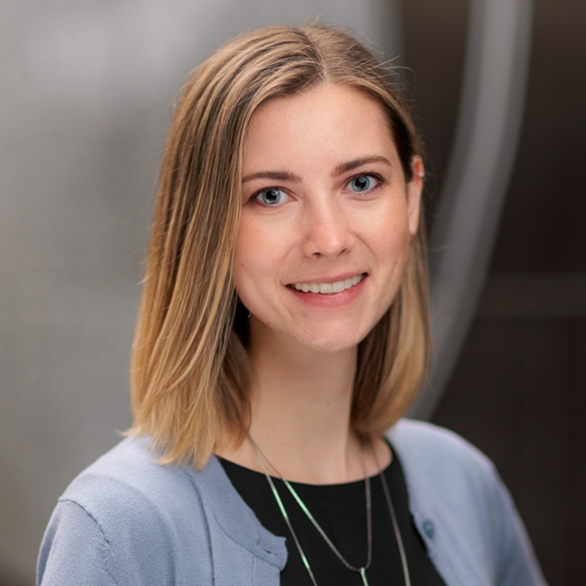 An image of a woman named Samantha Shumm. She is standing against a blurred gray background and smiling at the camera. She has light white skin, shoulder-length straight brownish blonde hair, and blue eyes. She is wearing a light blue cardigan over a black shirt.