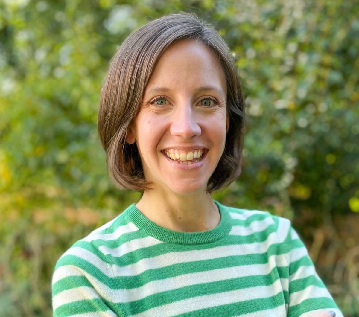 An image of a woman named Drea Braxmeier of the Milken Institute. She has white skin, bobbed brunette hair, and blue eyes. She is smiling at the camera, standing in front of greenery, and wearing a green and white striped shirt.