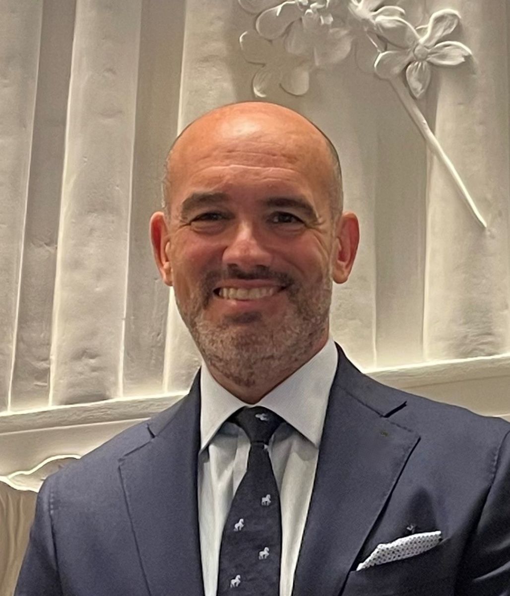 An image of a man named Manuel Martinez. He is smiling at the camera and has white skin, a bald head, and is wearing a navy blue suit with a tie.