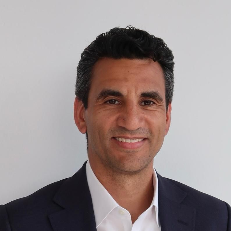 An image of a man named Ramon Escobar. He is smiling at the camera and has short dark brown hair with tan skin, dark eyebrows, and brown eyes. He is wearing a dark suit jacket over a white shirt and smiling at the camera against a white background. 