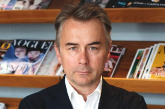 An image of a man named Albert Read. He is staring at the camera seriously against a background of a bookshelf with various magazines. He has white skin, short gray hair, and blue eyes and is wearing a dark blazer over a white collared shirt.