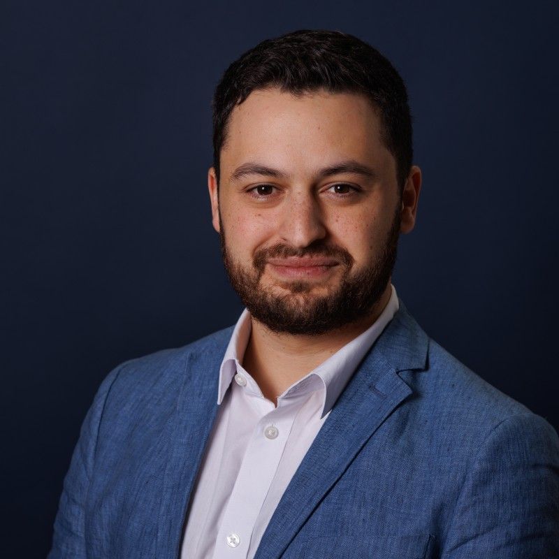 An image of a man named Ruben Turok. He has tan white skin, short black hair, a dark beard, and brown eyes. She is slightly smiling at the camera and standing against a dark blue background while wearing a lighter blue suit over a white shirt.