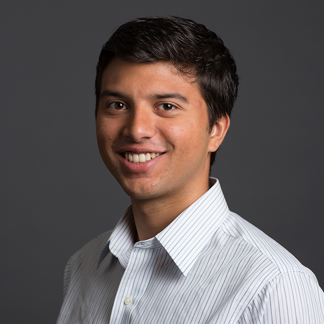An image of a man named Shiva Inalsingh. He has short black hair, tan skin, brown eyes, and he is wearing a white striped collared shirt and smiling at the camera against a dark gray background.