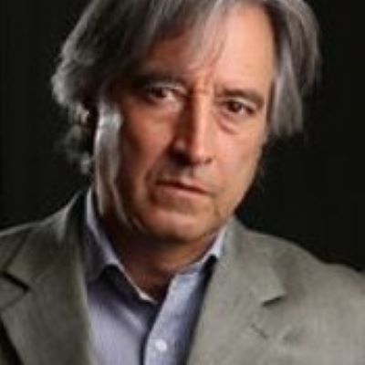 Adalberto Palma Gómez wearing a light gray blazer and blue shirt, looking seriously at the camera with a dark background.