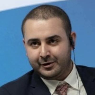 Adel Hamaizia speaking while wearing a dark suit and red tie, with an earpiece and a blue panel in the background.