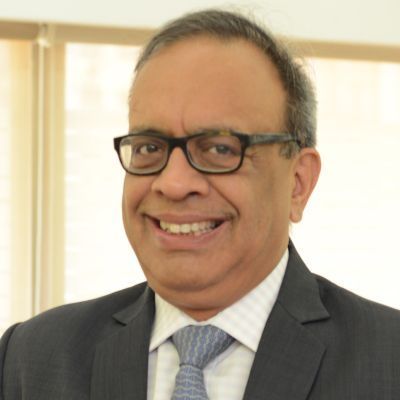 Alok Agarwal smiling in a suit and tie, wearing glasses, standing in front of softly lit blinds.