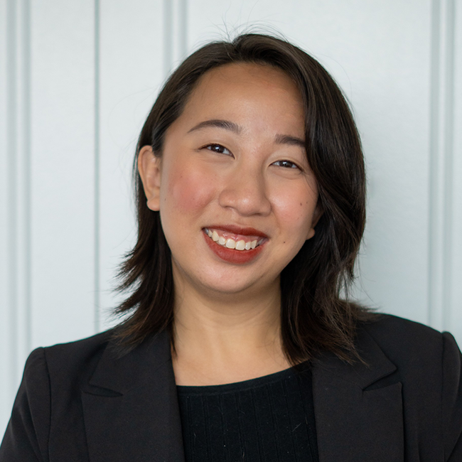 An image of woman named Ariana Salazar who is Asian in appearance with white skin and shoulder-length side-parted black hair. She is wearing a black blazer and standing against a white background and smiling. 