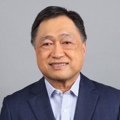 Cesar Purisima smiling in a navy suit and light blue shirt, with a plain gray background.