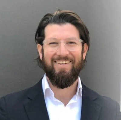 Enrique Perret Erhard smiling broadly in a dark suit and white shirt, with a full beard and clear glasses, standing against a neutral gray background.