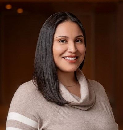 A woman named Gaby Alvarado smiles warmly while posing in front of a softly lit, blurred indoor background. She has long, straight dark hair and is wearing a light brown cowl-neck sweater with white stripes on the sleeves.