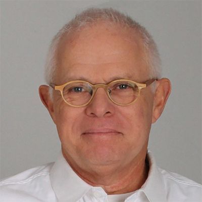 headshot of person with short cropped light hair, wearing gold round glasses and smiling subtly, wearing a whit collared shirt
