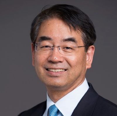 Heenam Choi wearing a suit and blue tie, smiling slightly in front of a dark gray background.