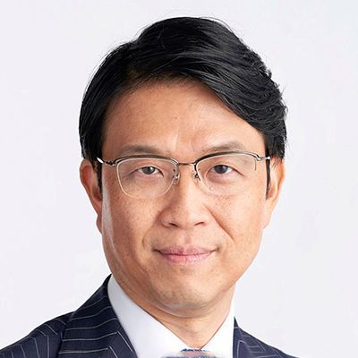 Hiro Mizuno wearing glasses and a pinstriped suit, looking directly at the camera with a composed expression against a light background.