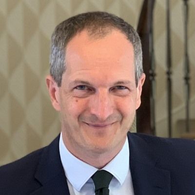 Lord James Bethell smiling in a suit and tie, standing indoors with a patterned background.