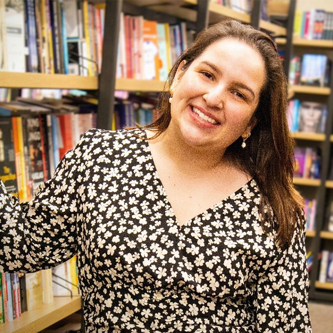 A woman named Michelle Rincon smiles warmly while standing in front of bookshelves in a bookstore or library. She is wearing a black dress with a white floral pattern and gold earrings, with her brown hair down and slightly parted to the side.