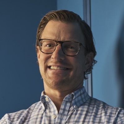 Stefan Haselwandter smiling in a checkered shirt and glasses, standing in front of a blue wall.