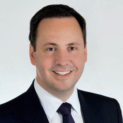 Steven Ciobo smiling in a dark suit and tie, with a white background."
