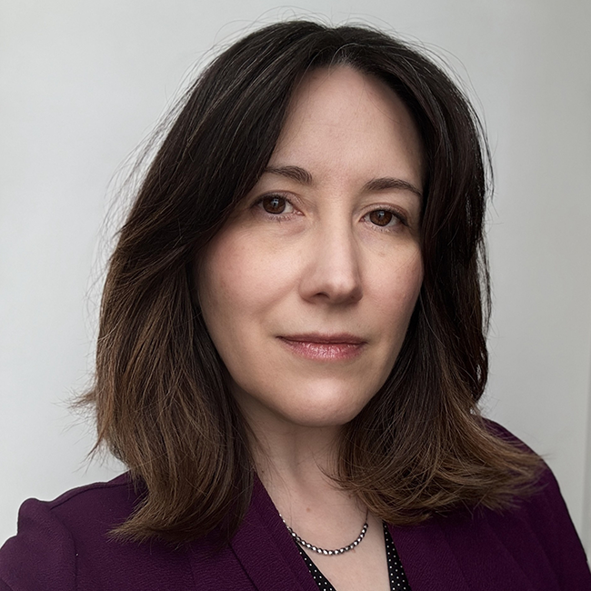 A woman named Susan Wetherby poses in front of a plain light gray background. She has straight, shoulder-length brown hair with subtle highlights and is wearing a purple blazer over a black top with small white polka dots. She has a calm, confident expression and is wearing a delicate beaded necklace.