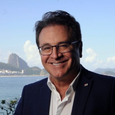 Vinicius René Lummertz smiling in a dark suit and glasses, standing in front of a scenic coastal backdrop in Rio de Janeiro.