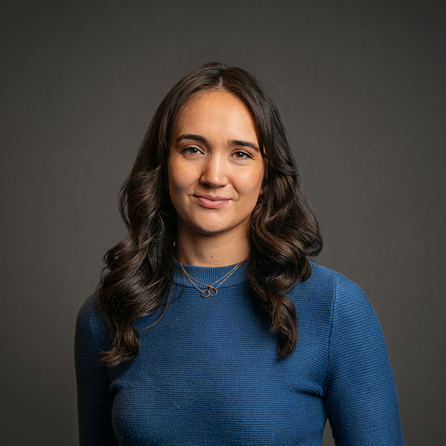 Professional headshot of Abigail Humphreys, a woman with long dark brown wavy hair, wearing a blue ribbed top and a gold necklace. She is smiling softly and posed against a dark gray background.
