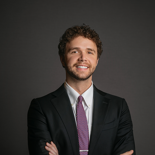 Professional headshot of Aidan Suski, a man with short curly brown hair and a beard, wearing a black suit with a white shirt and red-patterned tie. He is smiling confidently with arms crossed against a dark gray background.