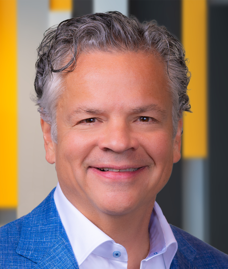 Alberto Casellas, a middle-aged man with short, wavy gray hair, wearing a blue textured blazer over a white dress shirt, smiling in front of a blurred background with yellow, gray, and black vertical elements.