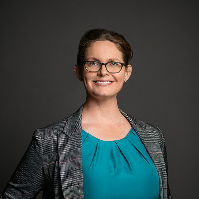 Professional headshot of Amanda Wagner, a woman with medium brown hair pulled back, wearing glasses, a teal top, and a gray plaid blazer. She is smiling and posed against a dark gray background.