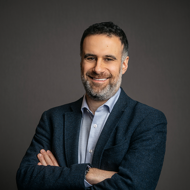 Professional headshot of Andrew Friedson, a man with short salt-and-pepper hair and a beard, wearing a navy blazer over a light blue button-down shirt. He is smiling with arms crossed, set against a dark gray background.