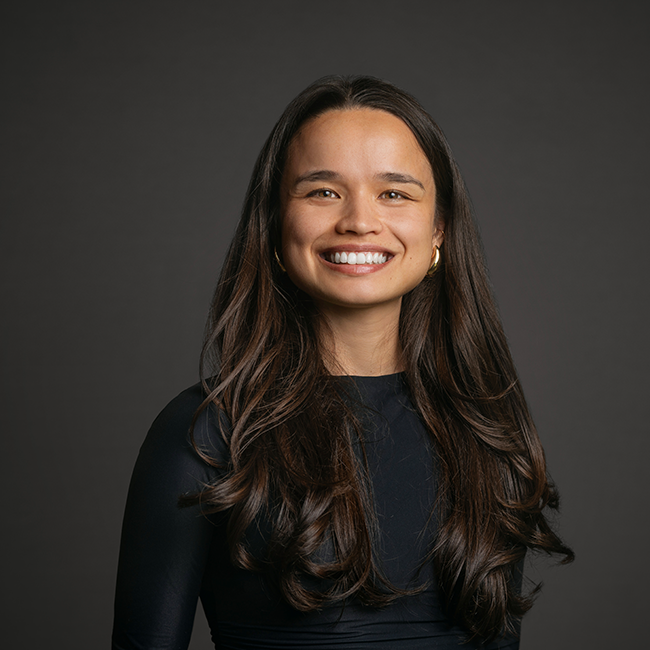 Professional headshot of Anna Lin-Schweitzer, a woman with long dark brown hair and a wide smile, wearing a navy top and gold hoop earrings. She is posed against a dark gray background.