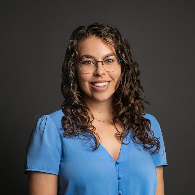Professional headshot of Athena Roesler, a woman with medium-length curly brown hair and glasses, wearing a light blue blouse with buttons. She is smiling and posed against a dark gray background.
