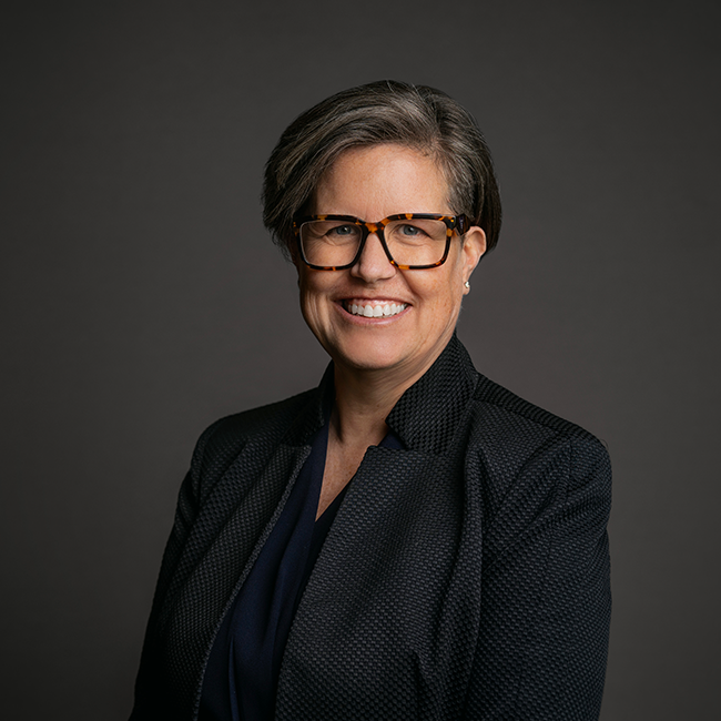 Professional headshot of Brenda Huneycutt, a woman with short gray hair and tortoiseshell glasses, wearing a dark textured blazer over a navy blouse. She is smiling broadly against a dark gray background.