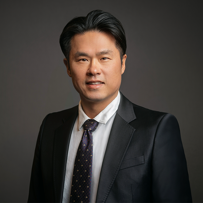 Professional headshot of Bumyang Kim, a man with neatly styled black hair, wearing a black suit jacket, white dress shirt, and a dark patterned tie. He is posed against a dark gray background with a neutral expression.