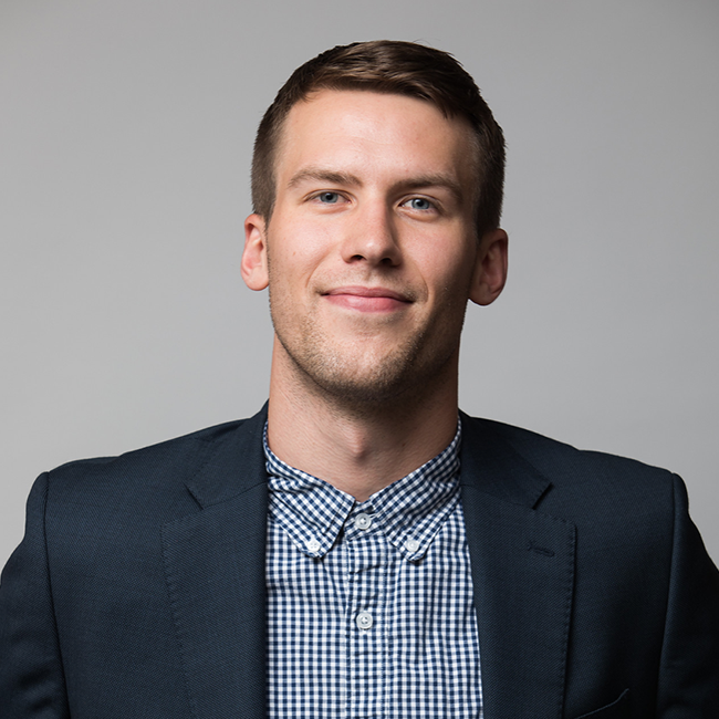 Professional headshot of Cameron McConkey, a man with short light brown hair, wearing a navy blazer over a blue and white checkered button-down shirt. He is smiling slightly and posed against a light gray background.