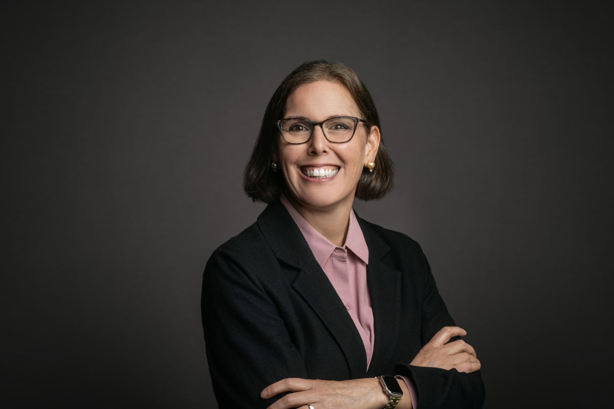 Professional headshot of Holly Freishtat, a woman with short brown hair wearing glasses, pearl earrings, a mauve collared shirt, and a black blazer. She is smiling with arms crossed and posed against a dark gray background.