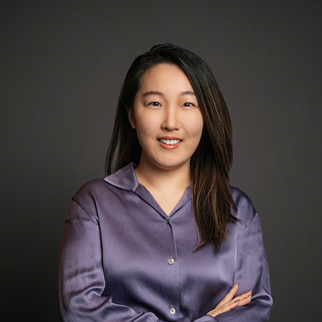Professional headshot of Jiayi Cullingford, a woman with straight dark brown hair and side part, wearing a purple satin blouse. She is smiling with arms crossed and posed against a dark gray background.