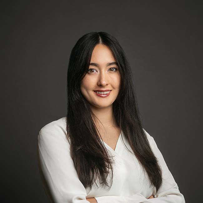 Professional headshot of Julia Hesse-Fong, a woman with long dark hair, wearing a white blouse and gold hoop earrings. She is smiling gently with arms crossed, set against a dark gray background.