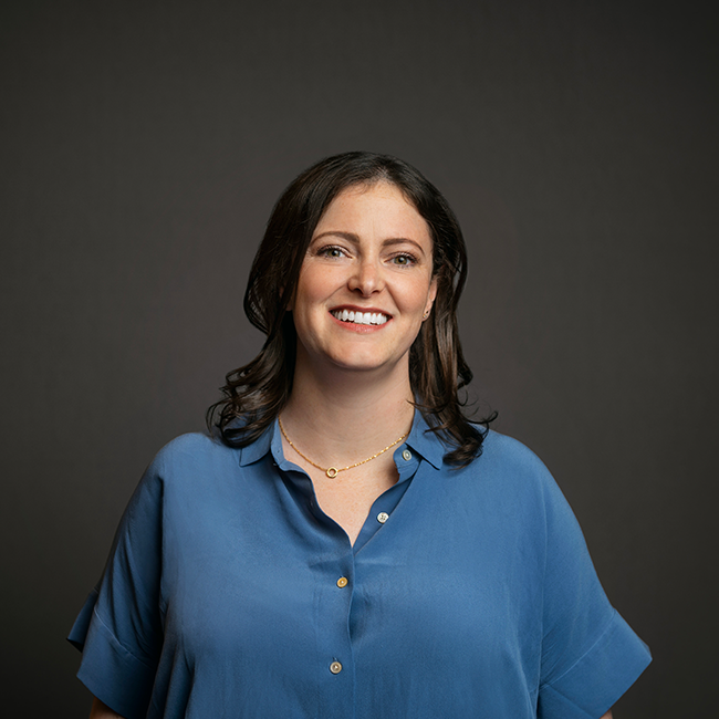 Professional headshot of Katherine Sacks, a woman with shoulder-length brown hair and soft curls, wearing a light blue blouse and a delicate gold necklace. She is smiling and posed against a dark gray background.